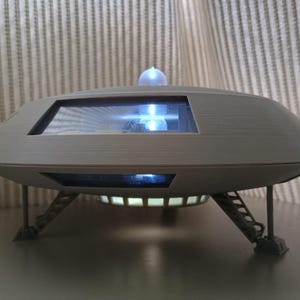 Puede incluir: Un objeto decorativo gris en forma de platillo volante con una ventana transparente y una pequeña cúpula brillante en la parte superior. El objeto tiene tres patas y una luz brillante debajo. El diseño sugiere un tema futurista o de ciencia ficción.
