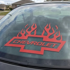Puede incluir: Un logotipo rojo de Chevrolet con llamas está adherido a la ventana trasera de un coche. El logotipo es una forma de corbata de lazo estilizada con la palabra "CHEVROLET". Las llamas son de color rojo brillante.