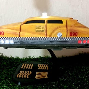 Puede incluir: Un modelo de taxi volador amarillo con borde a cuadros blanco y negro, marcas rojas B90 y una luz de techo blanca. El vehículo presenta un acabado desgastado, ventanas oscuras y un soporte negro con una base que muestra cuatro símbolos dorados.