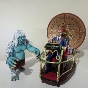 Op de afbeelding: Een blauw en wit figuur met lang wit haar staat naast een man in een blauw pak die op een gouden en rode stoel zit. De stoel is bevestigd aan een rond, gouden en rood platform met een grote, gouden schijf erachter.