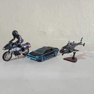 Puede incluir: Una motocicleta de juguete negra con un conductor, un coche deportivo de juguete negro y un helicóptero de juguete negro. El helicóptero tiene una pala de rotor blanca.