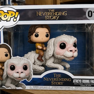 Peut inclure: Un ensemble de figurines en vinyle Funko Pop représentant Atreyu chevauchant Falkor de L'Histoire sans fin, dans une boîte de présentation. La boîte affiche le texte The Neverending Story, 01, et Atreyu Riding Falkor, avec une illustration des personnages.