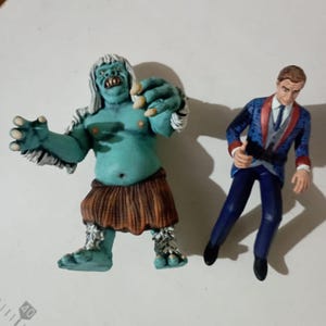 Op de afbeelding: Twee verzamelbare beeldjes. Eén is een turquoise huidmonster met wit haar en een bruine rok. De andere is een man in een blauw pak met een rode bies. Beide figuren staan op een wit oppervlak.
