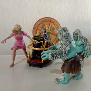 Op de afbeelding: Verzameling beeldjes die een scène uitbeelden met een vrouw in een roze jurk, een man in een stoel en twee blauwe wezens. De stoel heeft een grote, sierlijke, cirkelvormige achtergrond. De beeldjes staan op een wit oppervlak.
