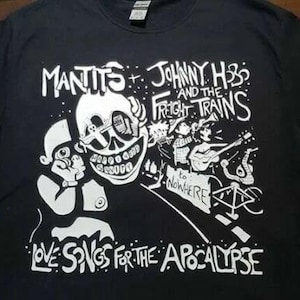May include: Black t-shirt featuring a white graphic design. The design includes the text "Mantits + Johnny H3 and the Freight Trains" and "Love Songs for the Apocalypse." The graphic depicts a skull, figures playing instruments, and the phrase "to Nowhere."