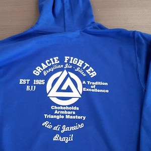 Puede incluir: Una sudadera con capucha azul real con el texto blanco "GRACIE FIGHTER" y "Brazilian Jiu-Jitsu". El diseño incluye un logotipo triangular, las palabras "EST 1925 BJJ", "A Tradition of Excellence" y "Rio de Janeiro Brazil".