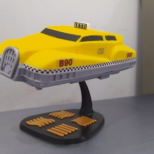 May include: A yellow and grey model taxi with the word "TAXI" on top. The model has the number "B90" on the side and a black and white checkered pattern along the bottom. It is mounted on a black stand with a unique base.