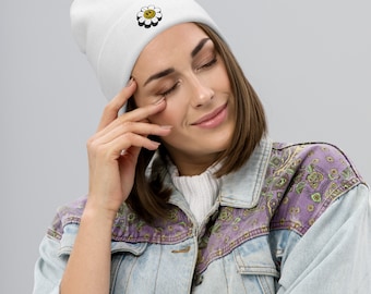 Puede incluir: Gorro blanco con una margarita negra y amarilla con una cara sonriente bordada. El gorro lo lleva una persona con el pelo castaño y una chaqueta vaquera azul claro.