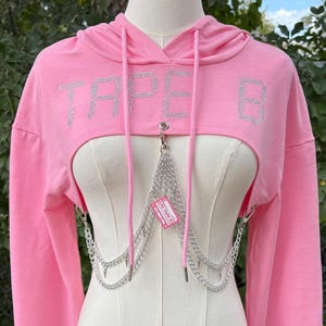 May include: A pink cropped hoodie with the word "TAPE B" in silver rhinestones across the chest. The hoodie features long sleeves and a drawstring hood. A silver chain harness is attached to the front.