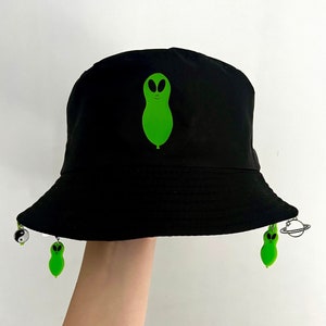 May include: Black bucket hat with a green alien graphic and two green alien charms attached. The hat has a wide brim and a flat crown.