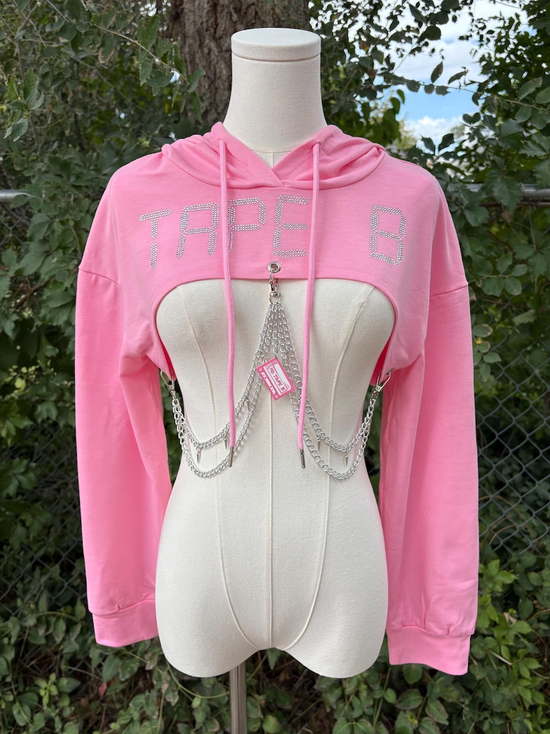 May include: A pink cropped hoodie with the word "TAPE B" in silver rhinestones across the chest. The hoodie features long sleeves and a drawstring hood. A silver chain harness is attached to the front.