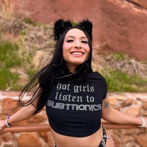 Pu&ograve; includere: Top corto nero con la scritta "hot girls listen to SUBTRONICS" in strass argentati. La persona indossa una fascia nera con orecchie soffici.