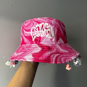 May include: Pink and white swirl-patterned bucket hat with the word "Scream" in white script. The hat has small charms hanging from the brim, including a cloud, mushroom, disco ball, and a rubber duck. The hat is held up against a gray background.