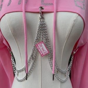 May include: Pink cropped hoodie with the word "TAPE" in rhinestone lettering. The hoodie features a silver chain detail with a small pink tag that reads "YO TAPE B". The hoodie has a drawstring and is displayed on a mannequin.