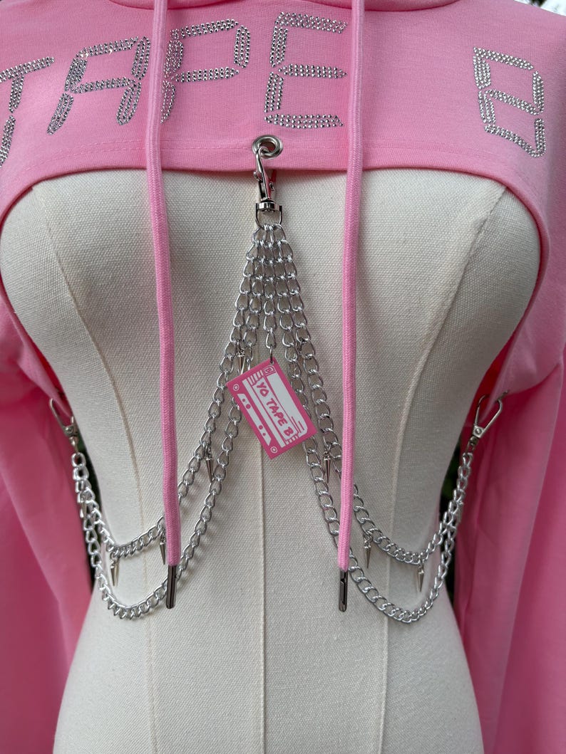 May include: Pink cropped hoodie with the word "TAPE" in rhinestone lettering. The hoodie features a silver chain detail with a small pink tag that reads "YO TAPE B". The hoodie has a drawstring and is displayed on a mannequin.