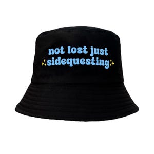 Könnte beinhalten: Schwarzer Bucket Hat mit dem Text "not lost just sidequesting" in Blau mit gelben Sternen auf beiden Seiten.