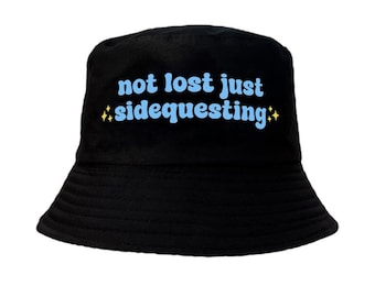 May include: Black bucket hat with the text "not lost just sidequesting" in blue with yellow stars on either side.