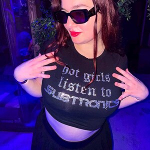 Pu&ograve; includere: Top corto nero con la scritta "Hot girls listen to SUBTRONICS" in strass argentati. Il top &egrave; abbinato a una gonna nera e occhiali da sole. L'immagine &egrave; scattata in un ambiente poco illuminato.