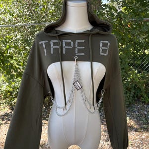 May include: An olive green cropped hoodie with long sleeves and a hood. The word "TAPE B" is spelled out in rhinestones across the chest. A silver chain with a small tag hangs from the neckline.