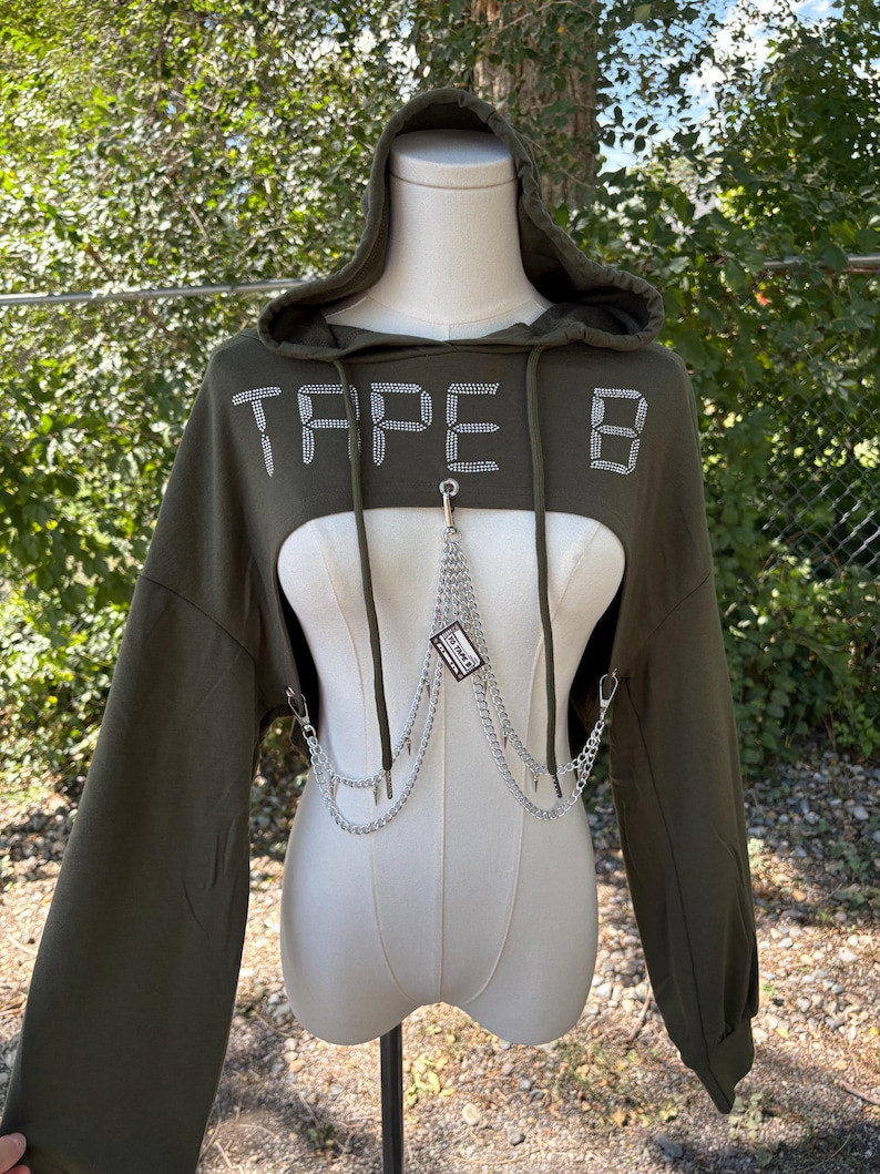 May include: An olive green cropped hoodie with long sleeves and a hood. The word "TAPE B" is spelled out in rhinestones across the chest. A silver chain with a small tag hangs from the neckline.