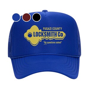 Op de afbeelding: Een koninklijk blauwe trucker-pet met een gele sleutelgrafiek en de tekst "Fugazi County Locksmith Co. "No questions asked."