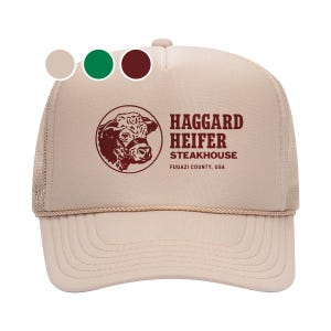 May include: A beige trucker hat with a brown mesh back. The front of the hat features a red logo with a bull's head and the text "Haggard Heifer Steakhouse, Fugazi County, USA."