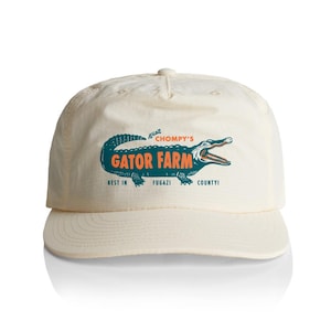 Puede incluir: Gorra de béisbol color crema con visera curva. La gorra presenta un gráfico de caimán verde azulado con las palabras "GATOR FARM" en naranja y el texto "BEST IN FUGAZI COUNTY!"