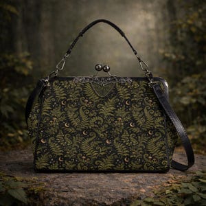 May include: A black handbag with a fern and moon pattern. The bag has a black handle and a detachable shoulder strap. The clasp is silver-toned with a decorative design. The bag is sitting on a stone surface.