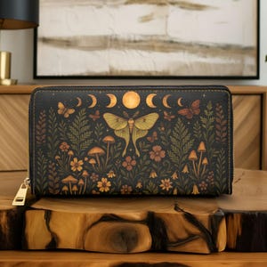 May include: Black zip-around wallet featuring a nature-inspired design. The wallet showcases a large moth, mushrooms, flowers, and crescent moons. The design is in shades of green, orange, and gold, with a gold zipper.