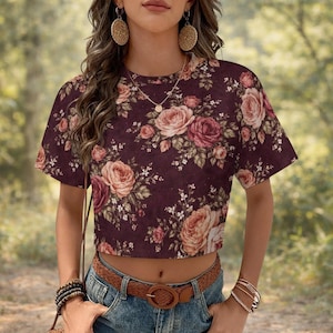 May include: A short-sleeve crop top in a dark burgundy hue featuring a dense floral pattern of pink, peach, and deep red roses with green leaves. The garment has a round neckline and is paired with blue denim jeans and a brown belt.