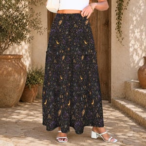 May include: A long, flowing skirt with a dark background and a celestial pattern. The design features crescent moons, stars, and floral elements in shades of purple, gold, and green. The skirt is worn with white sandals.