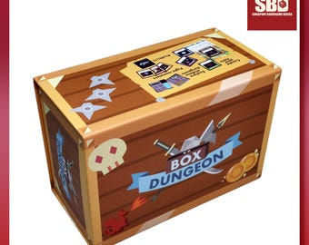 Pode incluir: Uma caixa de jogo de tabuleiro retangular castanha intitulada BOX DUNGEON com uma faixa azul clara, um ícone de caveira e ilustrações de armas. A tampa apresenta instruções e gráficos de cartas, o lado exibe o título do jogo.