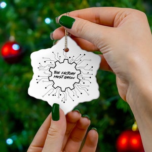May include: White snowflake-shaped Christmas ornament with the text "THE FACTORY MUST GROW" in a black, tech-inspired design. The ornament is held in front of a Christmas tree with red ornaments.