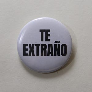 Può includere: Una spilla rotonda bianca con il testo TE EXTRAÑO stampato in grassetto nero a lettere maiuscole. La spilla ha una finitura liscia e lucida e un design semplice e pulito con il testo centrato sullo sfondo bianco.