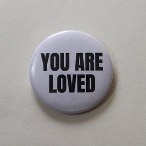 Può includere: Una spilla rotonda con sfondo bianco e il testo YOU ARE LOVED stampato in grassetto con lettere maiuscole nere. La spilla presenta una finitura liscia e lucida con un design semplice e pulito centrato sulla parte anteriore dell'oggetto.