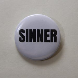 Può includere: Una spilla rotonda con fondo bianco e la parola SINNER stampata in lettere maiuscole nere, in grassetto e senza grazie, centrata sulla parte anteriore della spilla. Il design è semplice, pulito e ad alto contrasto, con una finitura liscia.