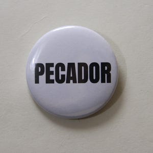Può includere: Una spilla rotonda con sfondo bianco e la parola PECADOR stampata in lettere maiuscole nere, in grassetto e senza grazie al centro. La spilla ha una finitura liscia e lucida e un design semplice e pulito per essere fissata agli indumenti.
