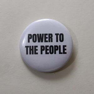 Può includere: Una spilla rotonda bianca con il testo nero POWER TO THE PEOPLE stampato in un carattere grassetto senza grazie al centro. La spilla presenta una finitura liscia e lucida, con un design semplice e pulito ad alto contrasto.