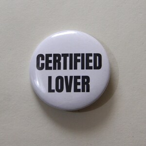 May include: A circular pin-back button with a glossy white surface. The text CERTIFIED LOVER is printed in bold, black, sans-serif capital letters across the center of the button. The design is simple, featuring a clean contrast between the black text and the white background.