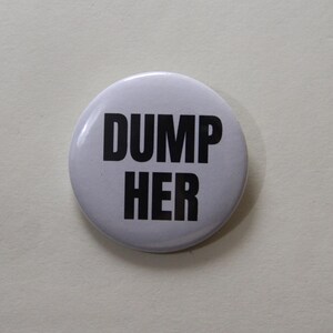 Könnte beinhalten: Ein runder, weißer Ansteckbutton mit dem Text DUMP HER in fetten, schwarzen Großbuchstaben, die mittig auf der Vorderseite platziert sind. Der Button hat eine glatte, glänzende Oberfläche und ein schlichtes Design mit hohem Kontrast.