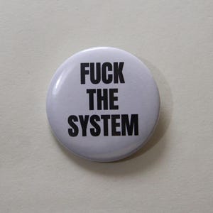 Può includere: Una spilla rotonda con sfondo bianco e testo nero in grassetto che recita FUCK THE SYSTEM. La spilla ha una finitura liscia e lucida e un design semplice, con il testo centrato su tre righe sulla superficie anteriore dell'oggetto.