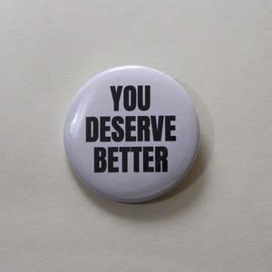 Può includere: Una spilla rotonda bianca con il testo YOU DESERVE BETTER stampato in lettere maiuscole nere, in grassetto e senza grazie. La spilla ha una finitura liscia e lucida, con un design semplice e un netto contrasto tra il testo nero e lo sfondo bianco.
