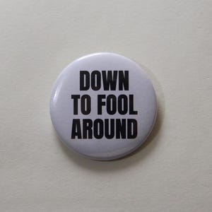 Può includere: Una spilla rotonda bianca con il testo DOWN TO FOOL AROUND stampato in grassetto nero a lettere maiuscole. La spilla ha una finitura liscia e lucida, con un design semplice e pulito centrato sulla parte anteriore dell'accessorio circolare.