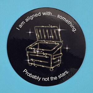May include: A round, black sticker with a gold outline of a treasure chest and the text "I am aligned with... something. Probably not the stars." The design includes star accents, creating a whimsical and humorous aesthetic.