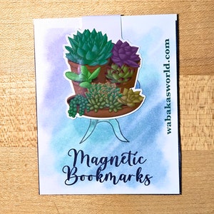 May include: A set of four succulent plants in terracotta pots, arranged on a tray. The plants are green and purple, and the tray is brown. The image has a watercolor background and the text "Magnetic Bookmarks" and "wahbakasworld.com".