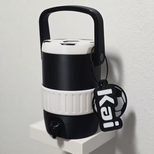 May include: A black and white water cooler with a soccer ball design on the lid. It has a black handle and a white band around the middle. A tag with the name "Kai" and a soccer ball hangs from the handle.
