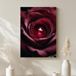 Op de afbeelding: Een ingelijste close-up van een dieprode roos met een gefacetteerde rode edelsteen in het midden. De rozenblaadjes zijn fluweelzacht en gedetailleerd, met een donkere achtergrond. Het kunstwerk wordt tentoongesteld op een lichtgekleurde muur, met witte vazen en een kopje op de voorgrond.