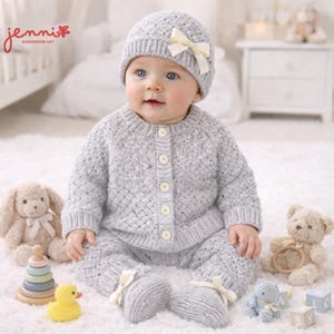 May include: A baby wearing a light gray knitted outfit, including a hat with a cream bow, a button-up cardigan, and matching pants with cream bows on the feet. The baby is surrounded by stuffed animals and toys, including a teddy bear and a wooden stacking toy.