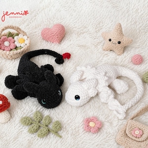 May include: A collection of handmade crochet items, including a black and a white dragon, a pink heart, a star, a basket of flowers, a mushroom, a leaf, a pink flower, a small purse, and a pink ball. The items are arranged on a white, fluffy surface.
