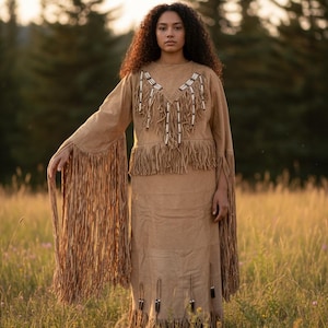 May include: A tan suede-like two-piece outfit with fringe and beaded accents. The top has long sleeves with fringe, and the skirt has fringe and decorative elements. The outfit is worn in a field of tall grass.